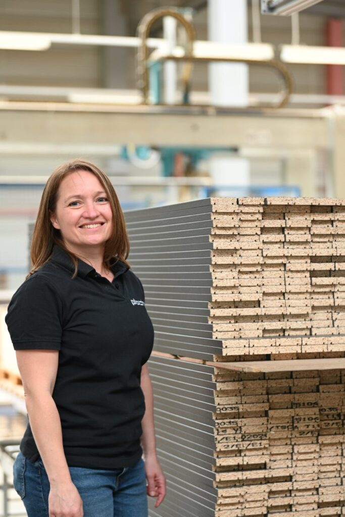 Une femme blonde, sans lunettes, se tient dans la production à côté d'une pile de profilés de matériaux finis.